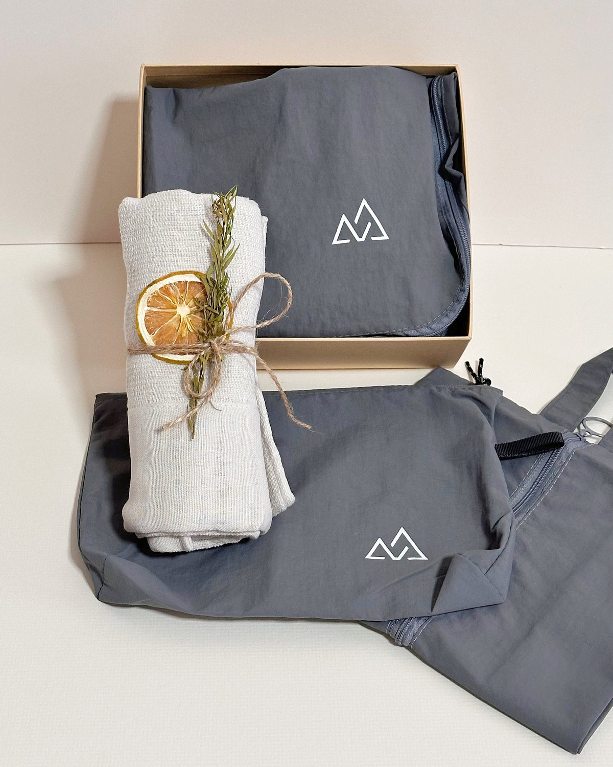 A folded gray fabric travel organizer sets item with a white mountain logo sits in an open kraft gift box, alongside a rolled off-white fabric cotton towel item tied with twine and a dried orange slice, on a light beige background.