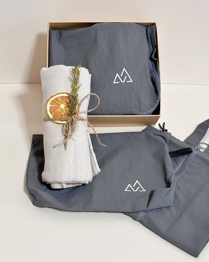 A folded gray fabric travel organizer sets item with a white mountain logo sits in an open kraft gift box, alongside a rolled off-white fabric cotton towel item tied with twine and a dried orange slice, on a light beige background.