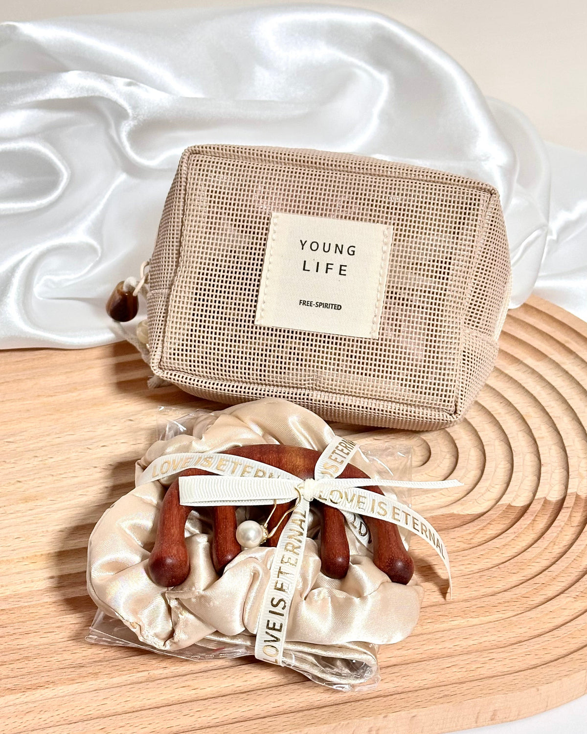 A champagne or beige mesh zip pouch with a cream label reading 'YOUNG LIFE' sits on a wooden board beside a satin scrunchie with wooden claw clips and a matching ribbon.