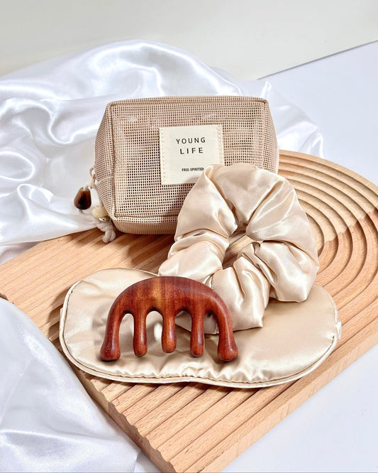 A hair care gift set featuring a beige mesh pouch with a 'YOUNG LIFE' label, a beige satin scrunchie, a champagne or beige satin eye mask, and a reddish-brown wooden hair massager, arranged on a wooden tray with white satin fabric.