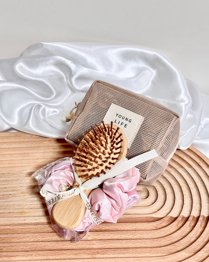 A hair care set featuring a wooden hairbrush with brown bristles, a beige mesh pouch labeled 'YOUNG LIFE', and one pink scrunchy and one satin eye mask wrapped in clear plastic with a ribbon, arranged on a wooden tray with white satin fabric in the background.