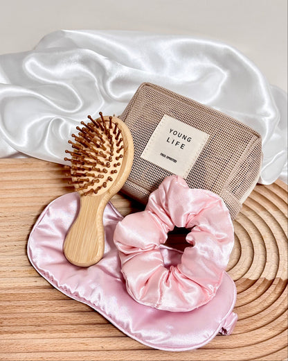 Wooden hairbrush, pink scrunchie and eye sleeping mask, and brown pouch on a wooden surface with white fabric in the background.